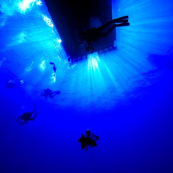 Ein Tauchboot ist von unten aus der Tiefe gegen das Sonnenlicht fotografiert. Alles in einem tiefen Blau. Taucher sind in der Silhouette zu erkennen