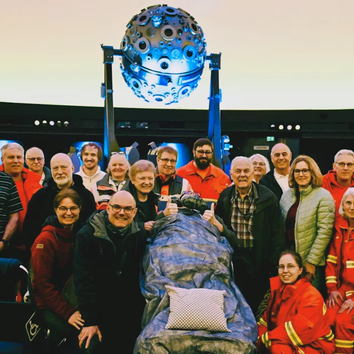 Eine Gruppe von Freunden und Mitarbeiter:innen der DRK stehen um einen Patienten im Transportbett vor dem Universarium im Planetarium Stuttgart