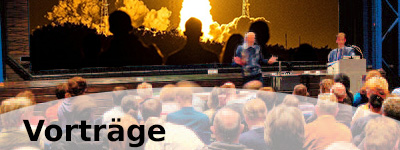 Bild zeigt zwei Menschen auf einer Bühne, ein Raketenstart im HIntergrund und viele Menschen (Blick von hinten) sitzen im Publikum