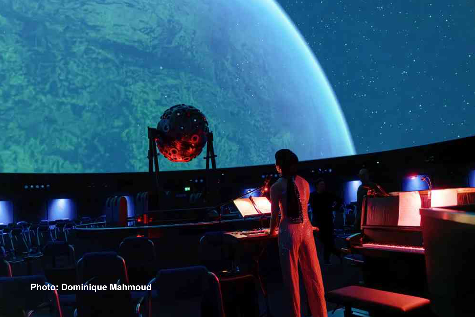Eine Frau steht mit Notenblatt in der Mitte des Bildes, spielt auf einem Synthesizer. Im Hintergrund ist das Universarium zu sehen, dass rot angestrahlt ist.