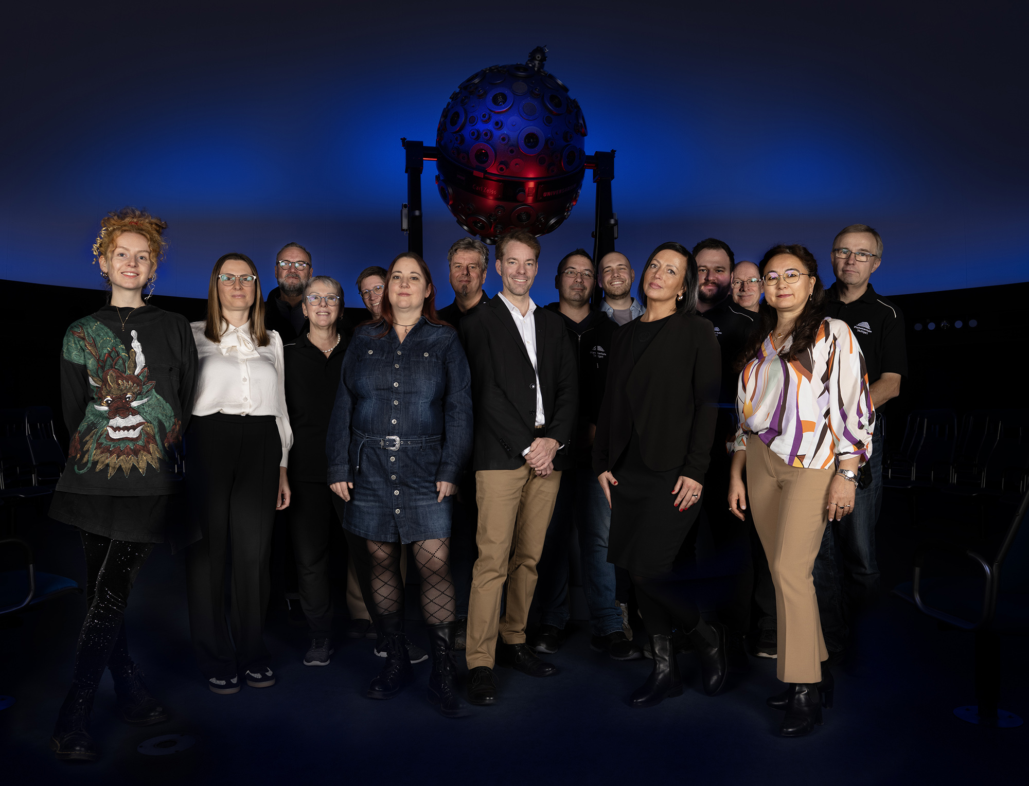 15 Kolleg:innen aus dem Planetarium Team stehen als Gruppe in der dunkelblau erleuchteten Sternenkuppel vor dem Universarium.