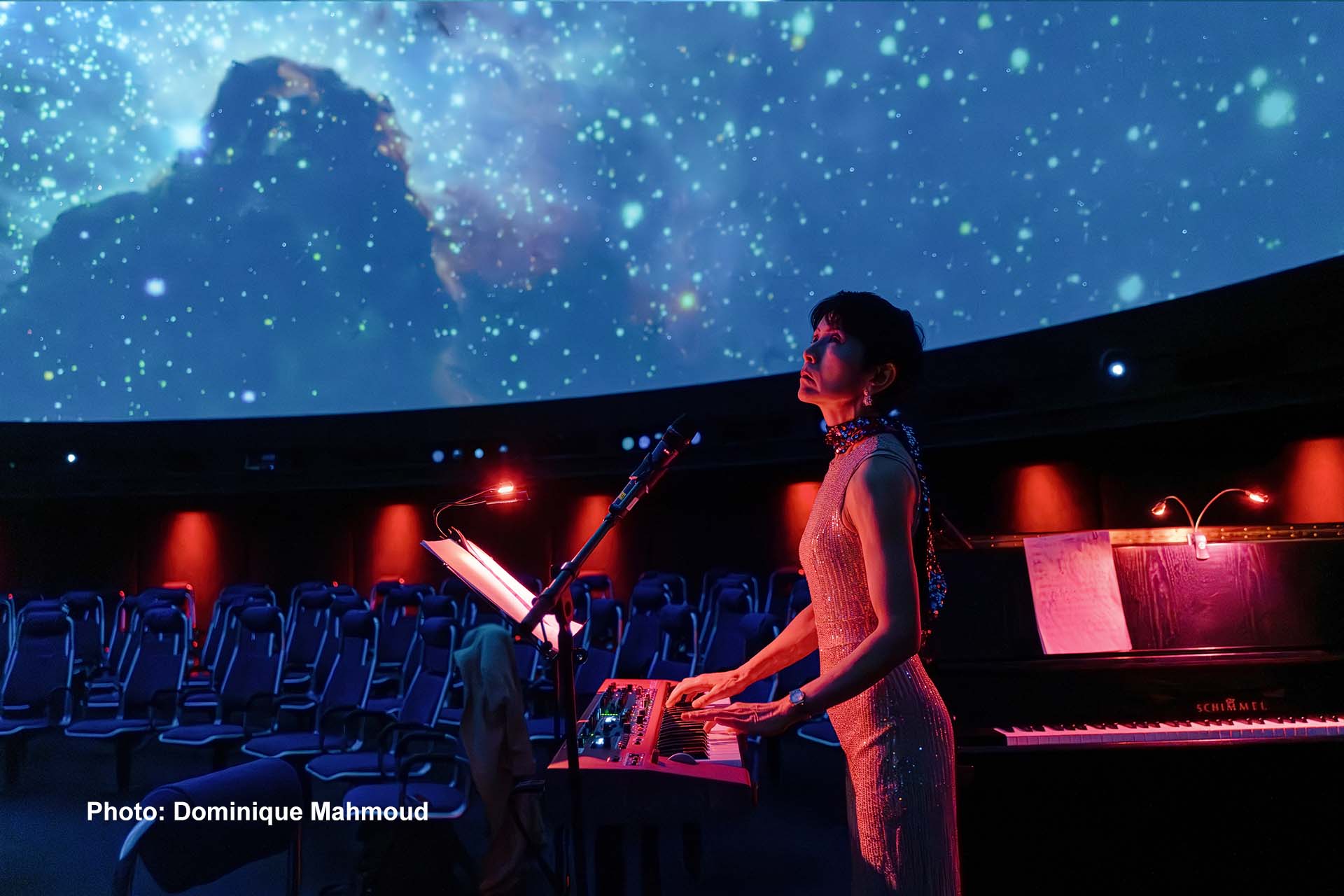 Eine Frau steht an einem Synthesizer, rot angeleuchtet. Im Hintergrund ist der Sternenhimmel im Planetarium zu sehen.